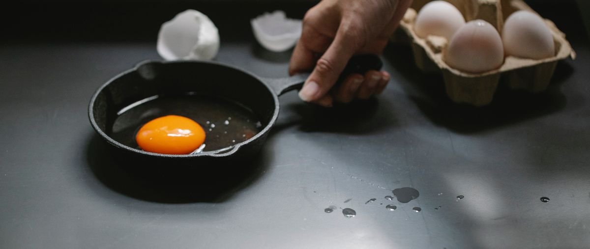 Fresh egg cooking in a small black skillet next to a carton of eggs