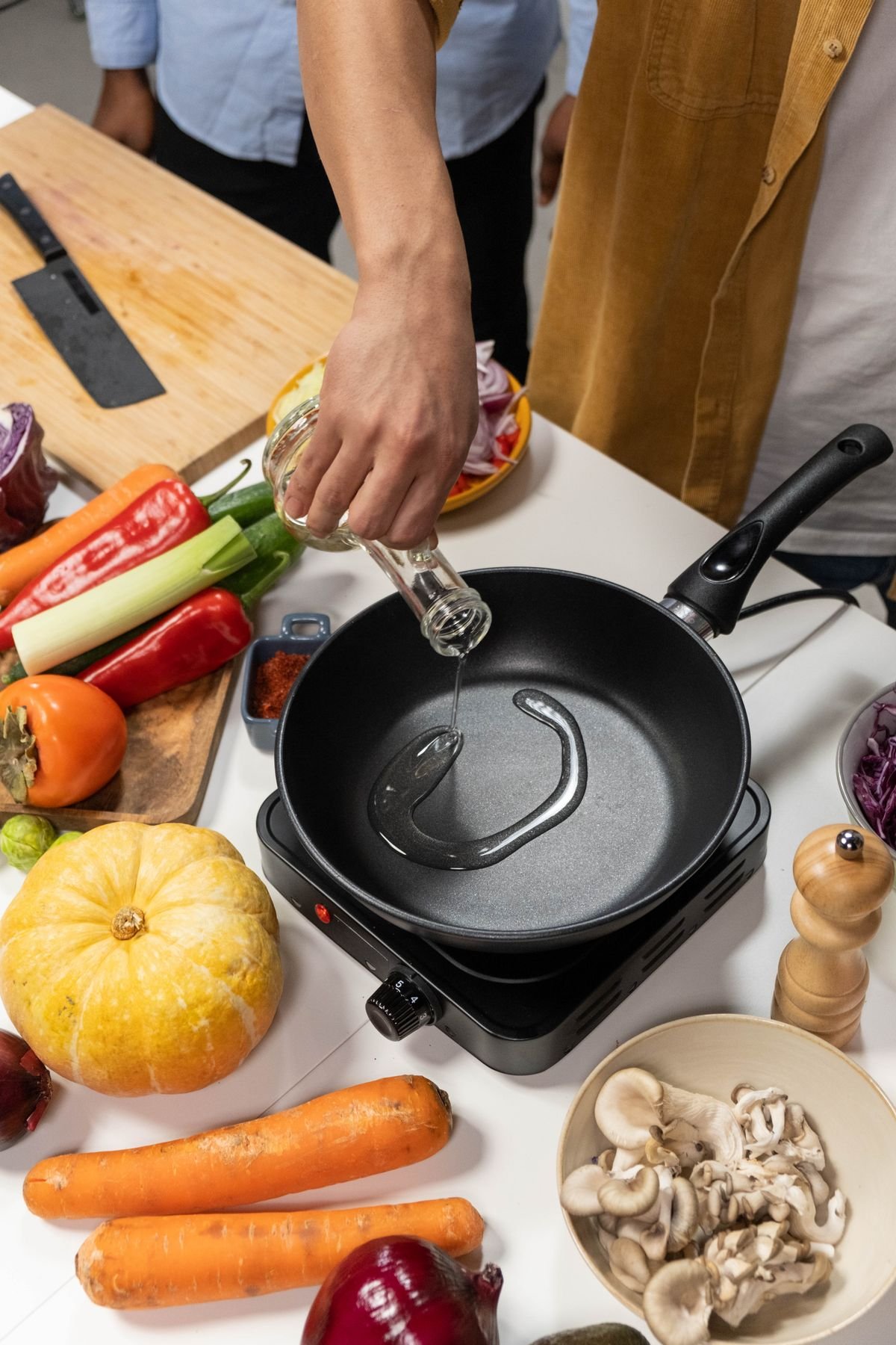 Hand pouring a thin stream of cooking oil into a dark pan on a hot plate